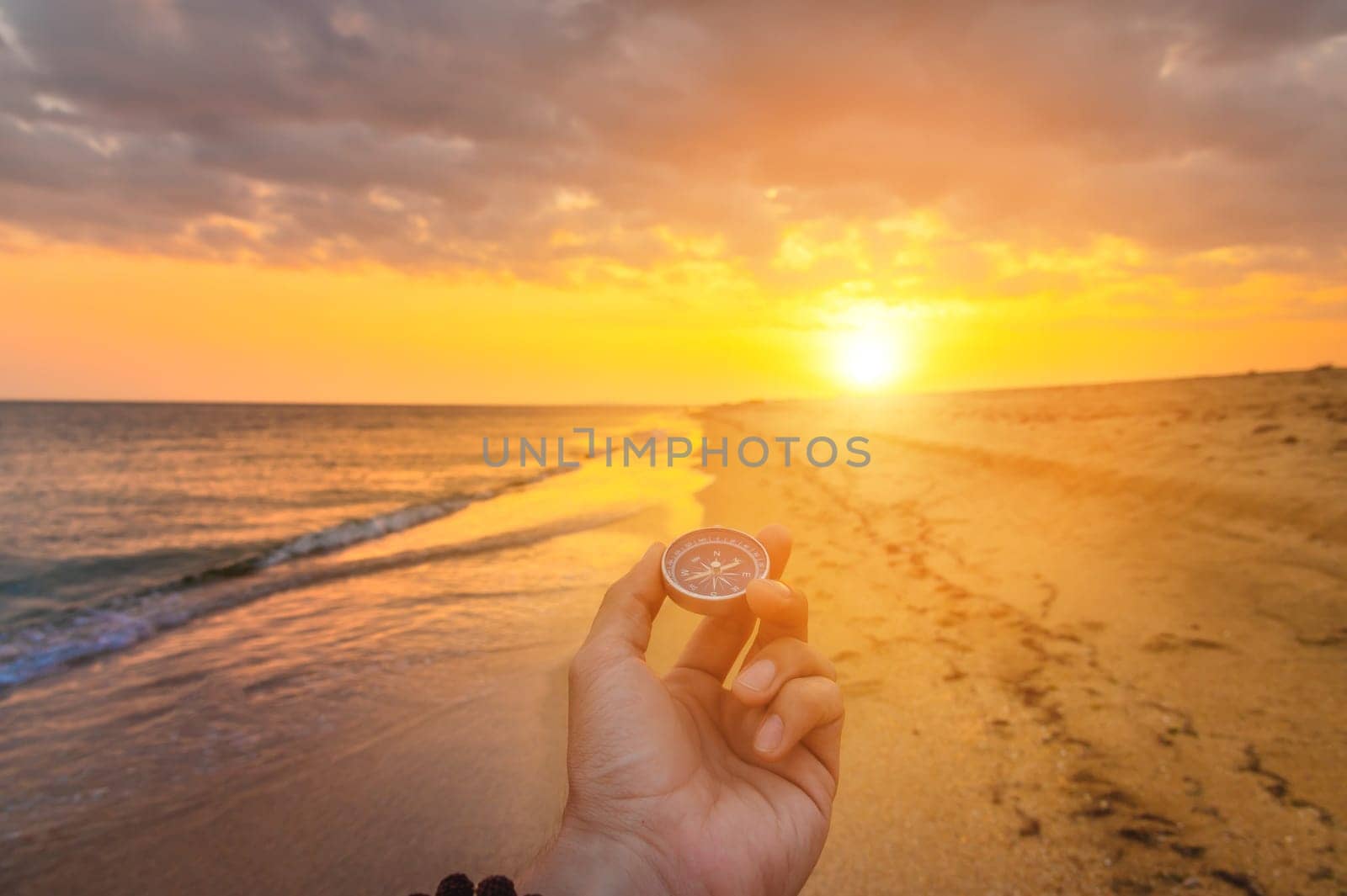 First person view of a man's hand with a compass against the backdrop of a beautiful seascape. Navigation concept of finding your way and orientation. The setting sun with rays enters the frame.