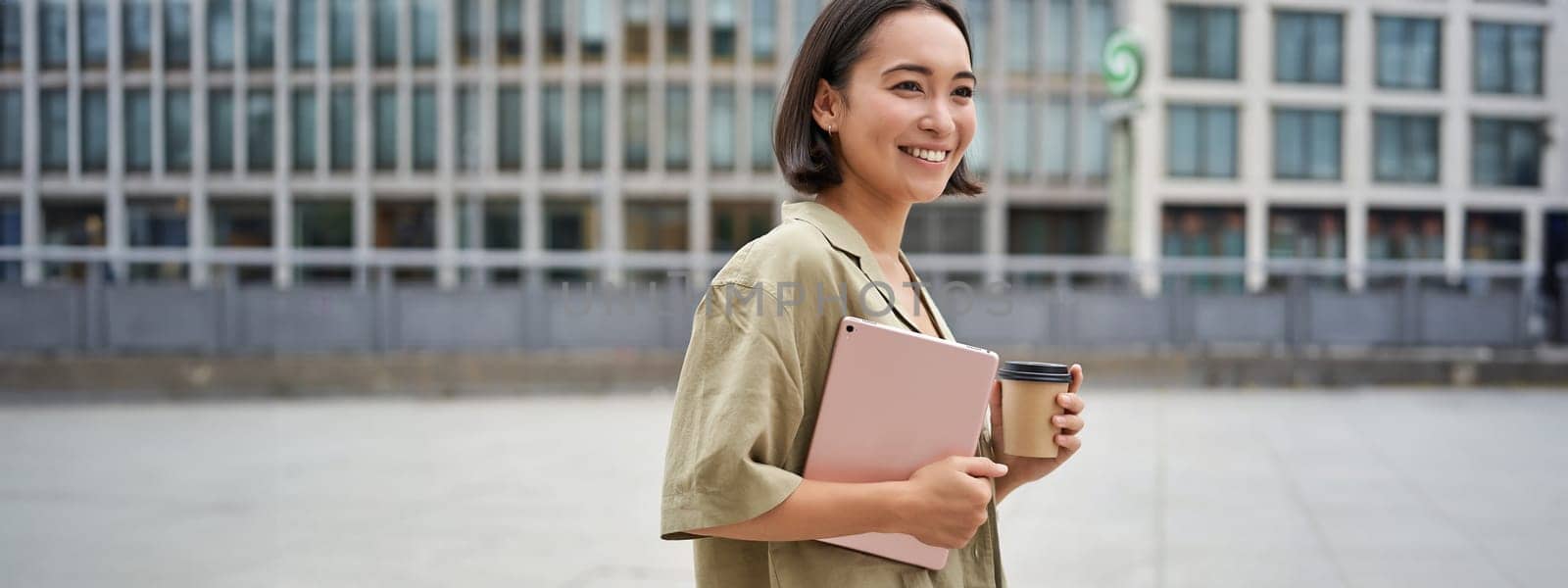 Portrait of happy asian girl with tablet and coffee, walking on street with pleased smile.