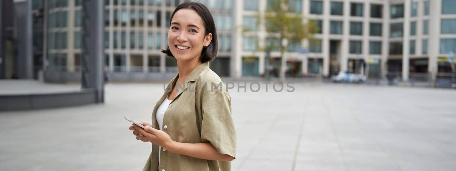 Portrait of asian woman standing on street, city square and holding mobile phone. Girl with smartphone walking outdoors.