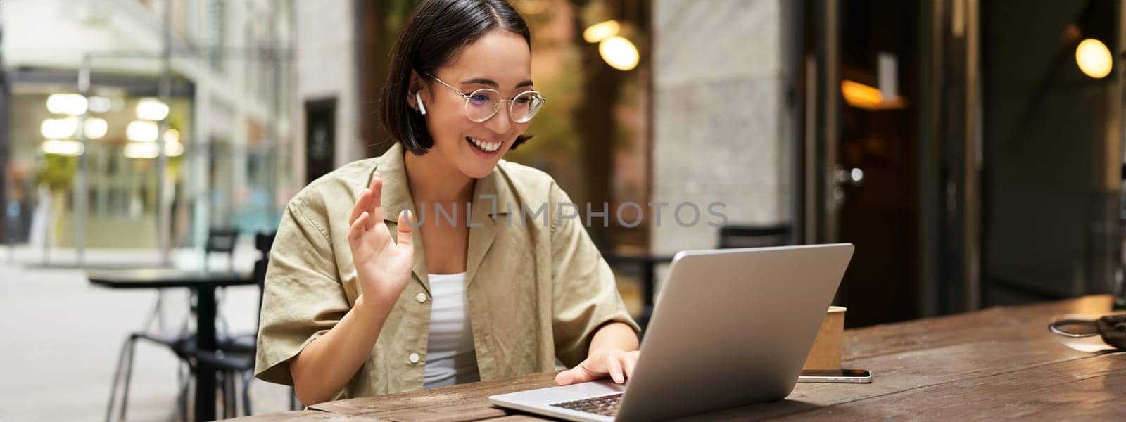 Online meeting. Young urban girl, asian woman talking with laptop, video chat, gesturing, sitting in an outdoor cafe and working remotely by Benzoix