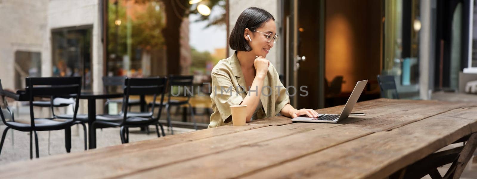 Silhouette of young woman working in cafe with laptop, typing on keyboard and drinking coffee, sitting outside.