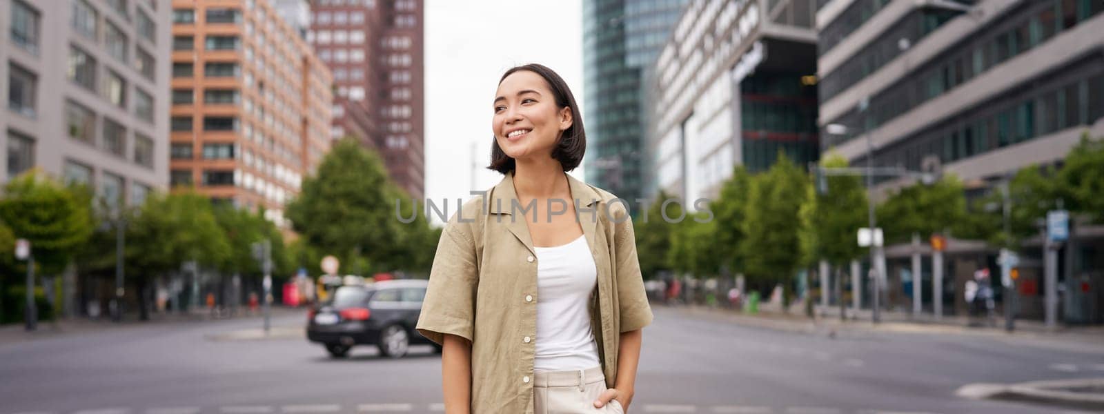 Outdoor shot of young smiling asian woman, standing on street in daytime, looking around, posing happy by Benzoix