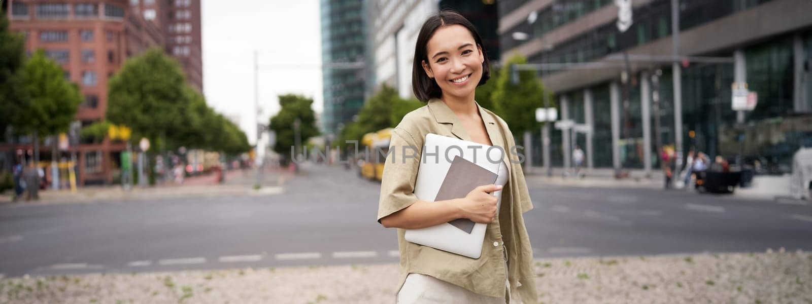 Portrait of young asian woman, looking happy and confident, going to work or university, city skyscrappers behind her, holding laptop and notebook by Benzoix