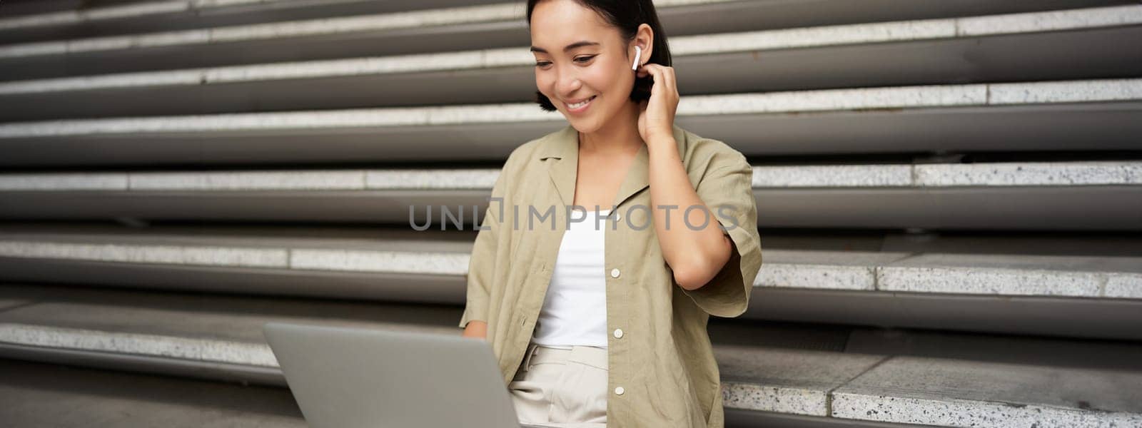 Remote worker. Smiling asian girl sits outdoors on street with laptop. Happy young woman working on computer remotely, listen music in wireless headphones by Benzoix