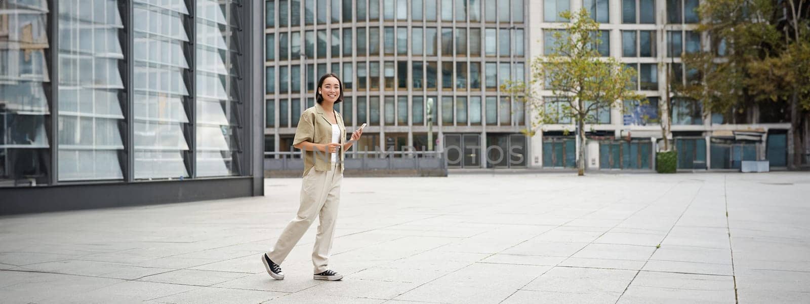 Portrait of beautiful smiling asian woman, walking on street of city centre with smartphone, looking at camera by Benzoix