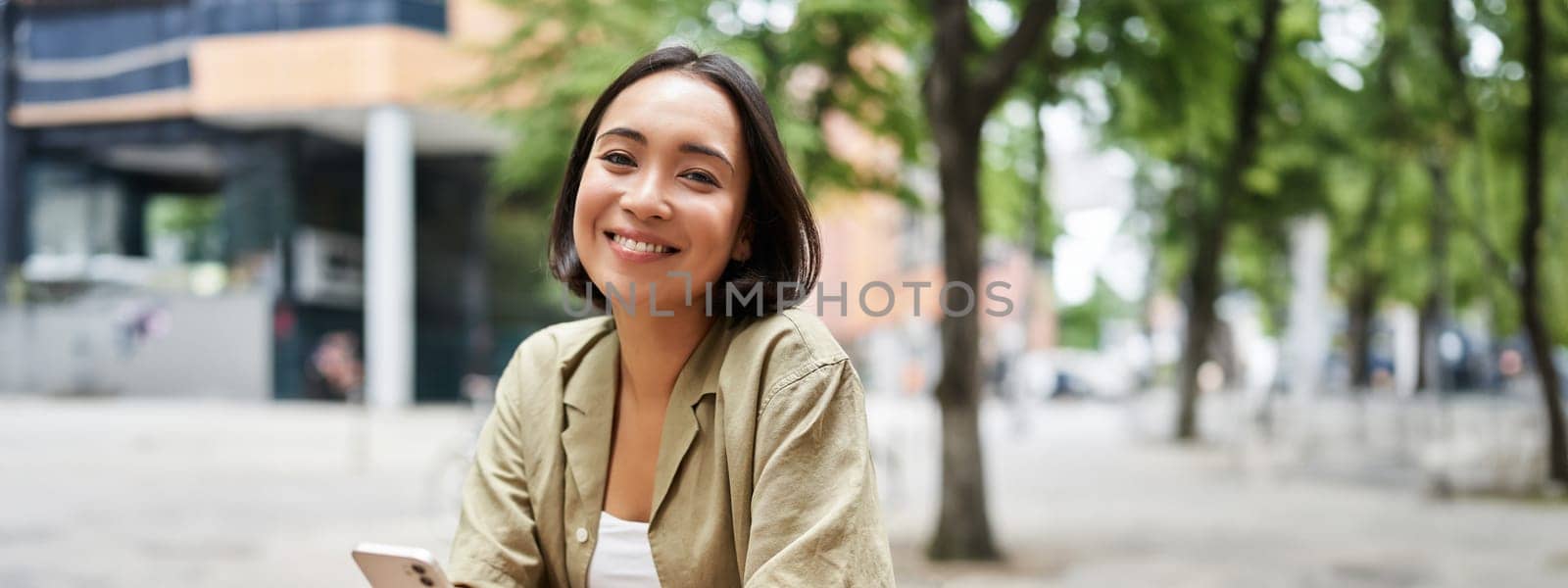 Portrait of young woman enjoying her coffee, drinking takeaway on bench in city, using smartphone by Benzoix