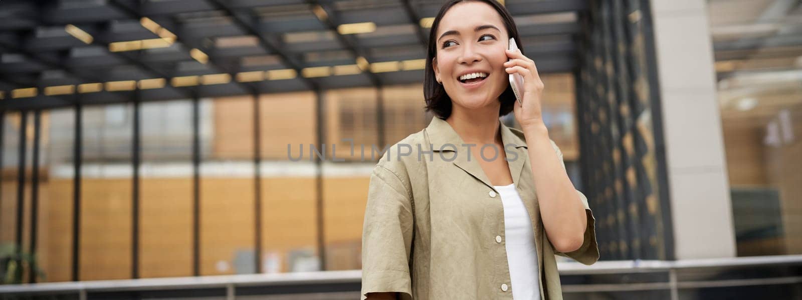 Happy asian woman talking on mobile phone, standing on bus stop in city. Outdoor shot.