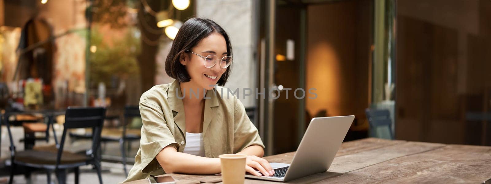 Young woman working in a cafe, using laptop and drinking coffee. Asian girl student with computer studying remotely, sitting on bench near shop.