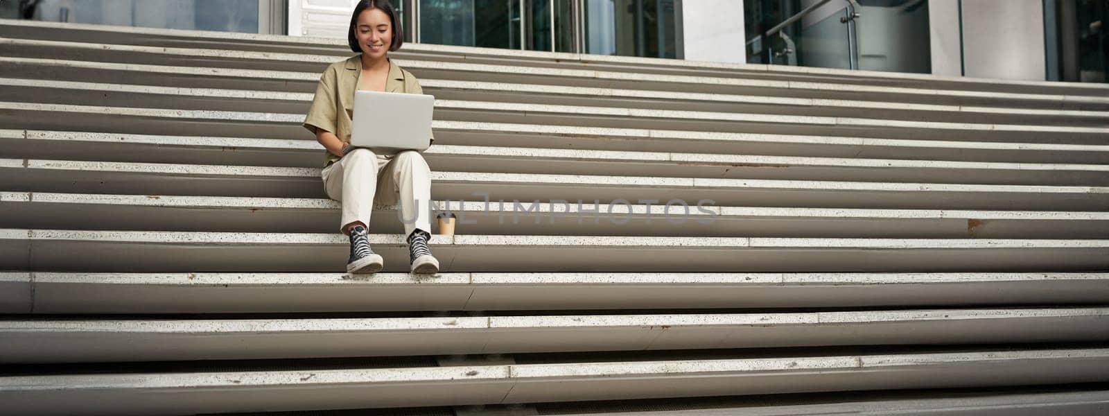 Asian girl student sits on stairs near campus, types on laptop, does her homework outdoors by Benzoix