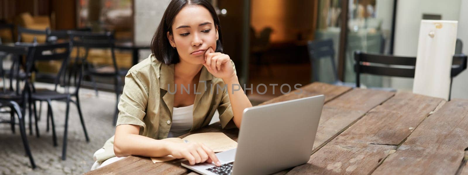 Bored girl looks at her laptop, sits outdoors in cafe, listening boring online meeting, working and feeling sad by Benzoix