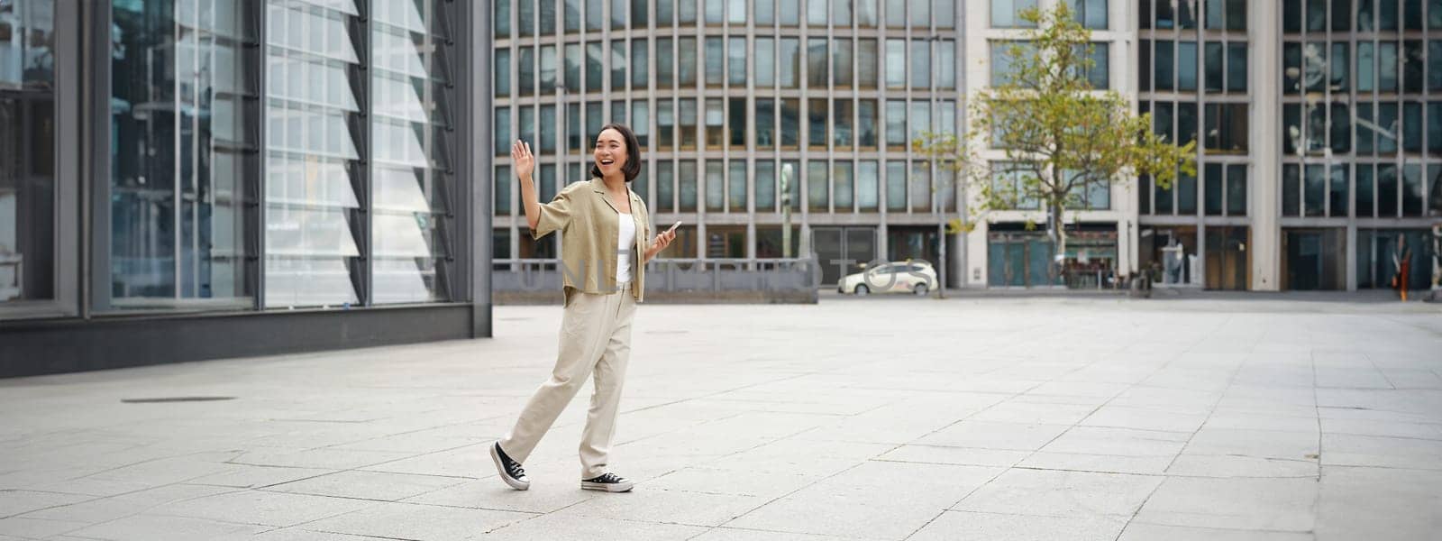 Happy asian girl saying bye to someone, waving at friend and walking on street with smartphone.