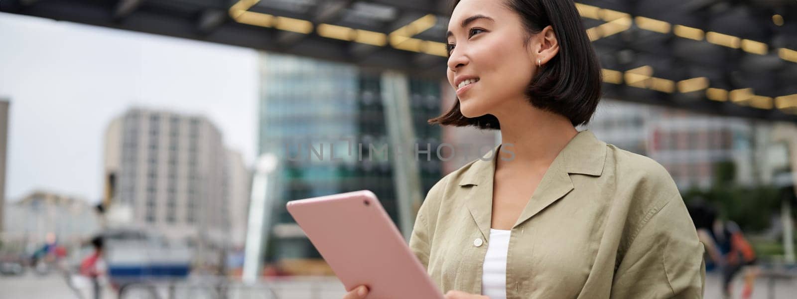 Portrait of asian woman standing on street, using tablet, smiling with carefree face expression, outdoor shot.
