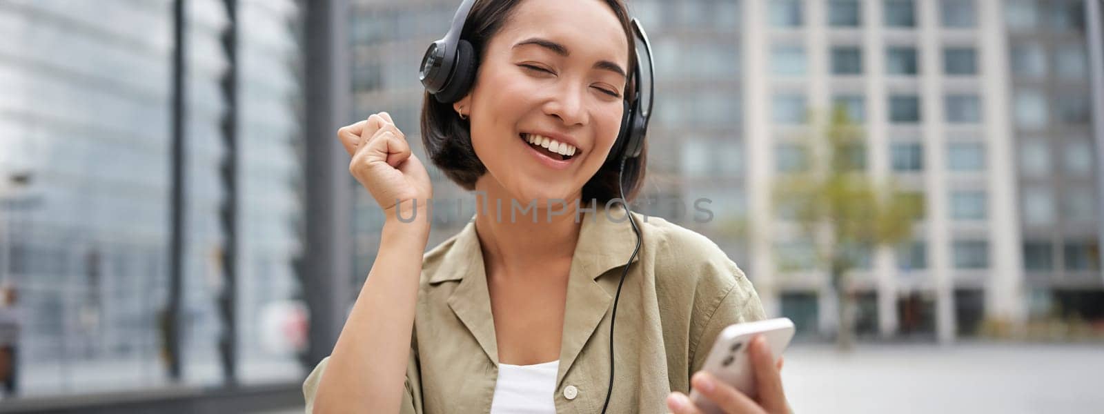 Happy girl dances on street and listens music in wireless headphones, holds mobile phone, using streaming app by Benzoix