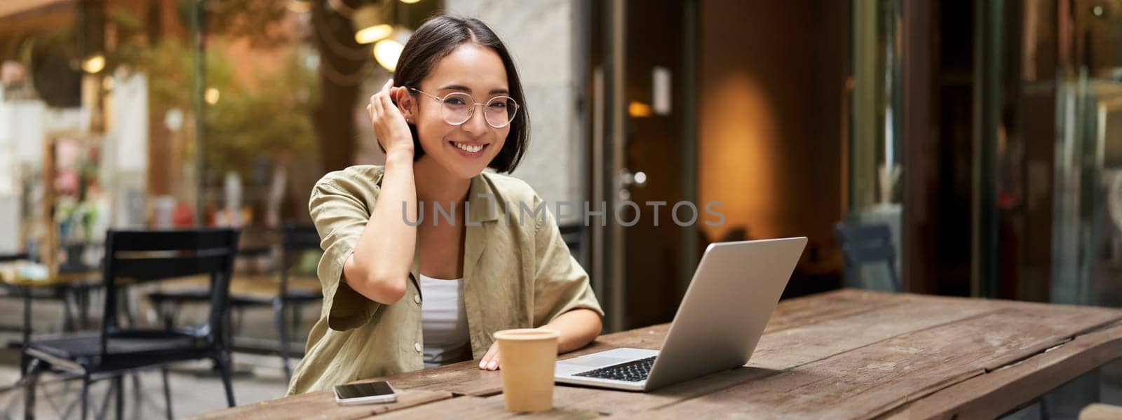 Portrait of smiling girl in glasses, sitting with laptop in outdoor cafe, drinking coffee and working remotely, studying online.