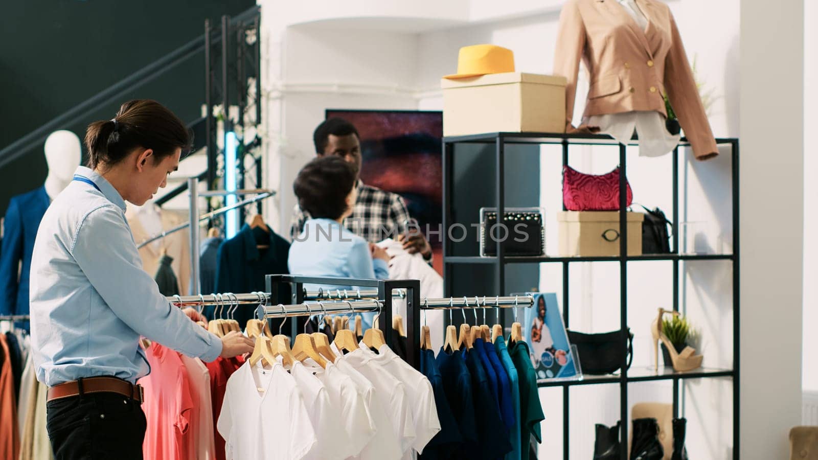 African american customer asking worker for help in modern boutique, shopping for stylish merchandise. Asian employee arranging hangers with fashionable clothes in clothing store. Fashion concept