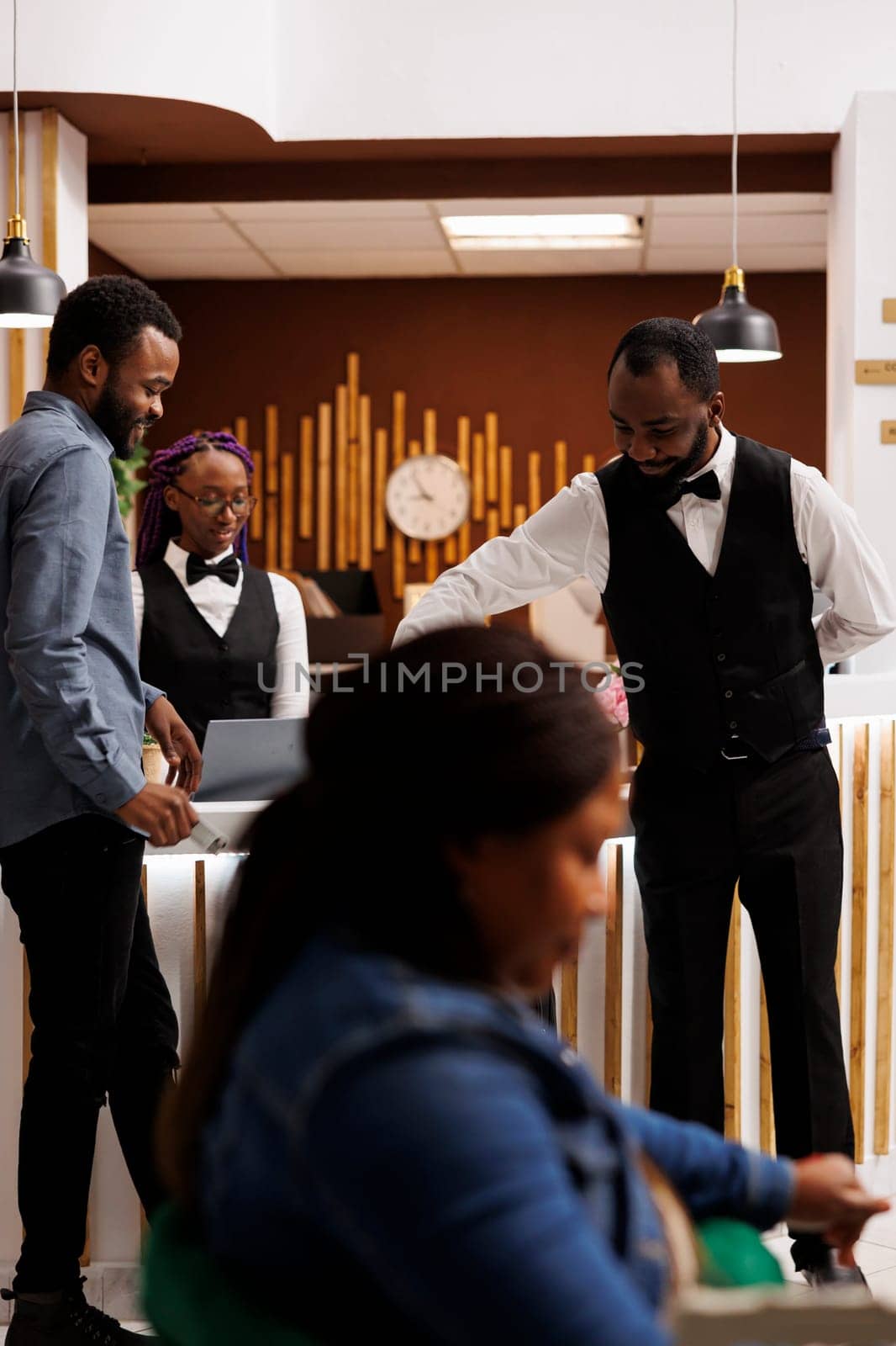 Young African American smiling guy bellboy providing customer service to hotel guest, moving his luggage, carrying suitcase to room after check-in. Bellhop and tourist communicating at front desk