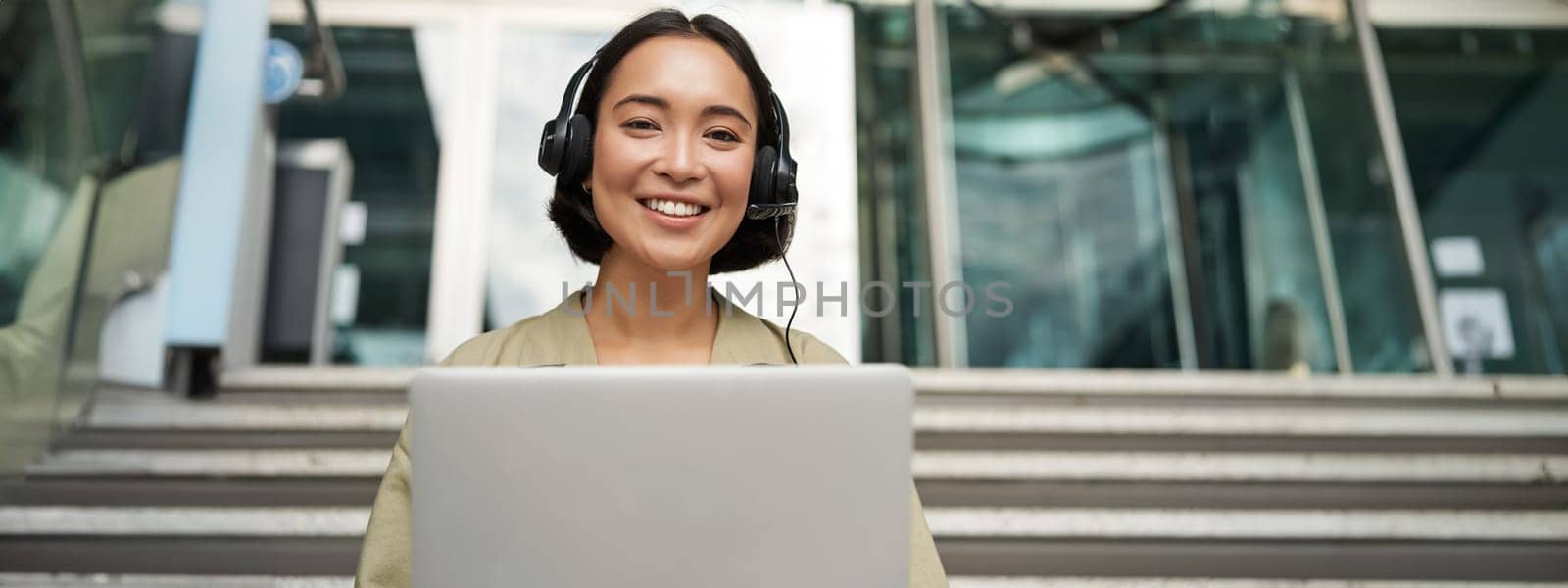 Portrait of young asian woman sitting with laptop and headphones, watching video, does online course on computer, sitting on stairs outdoors.