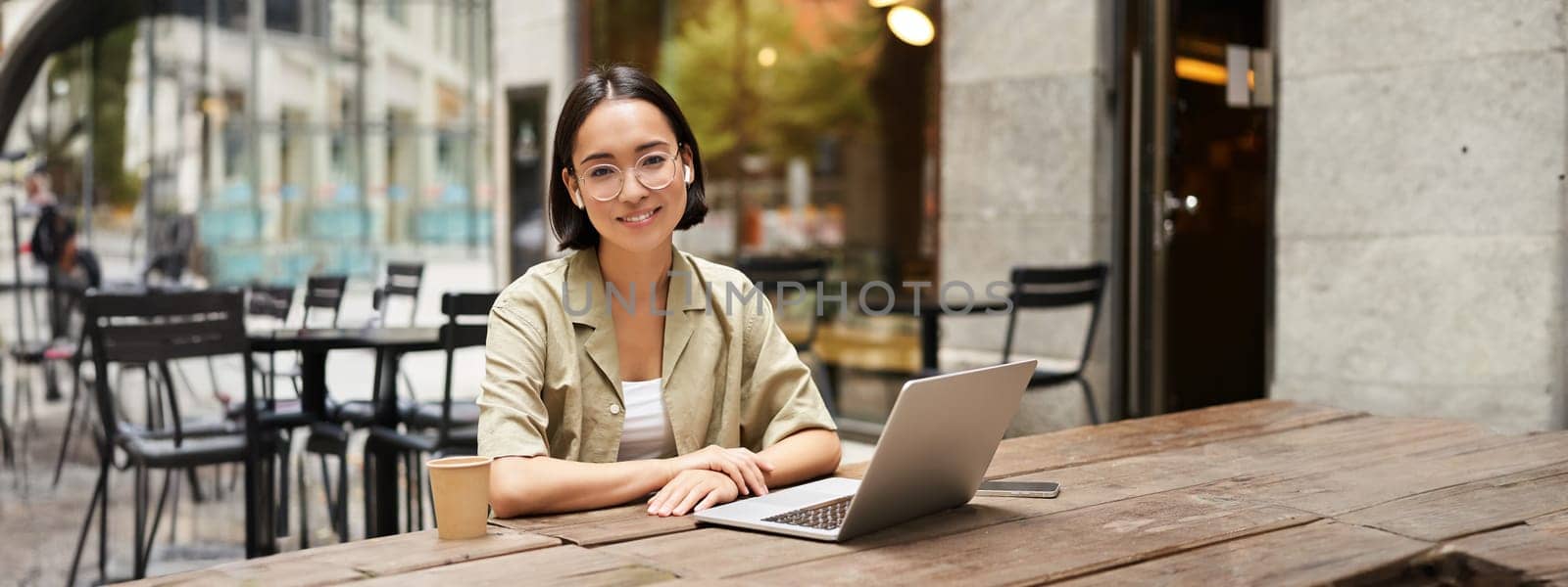 Young woman, student working from cafe, sitting with laptop and cup of coffee outdoors, looking at camera by Benzoix