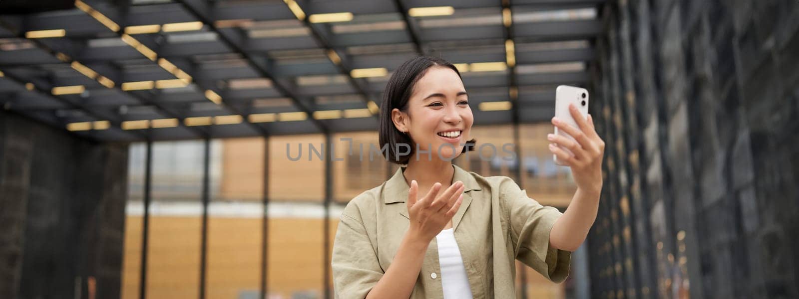 Happy asian girl shows something behind her during video call, demonstrating smth, standing on street by Benzoix