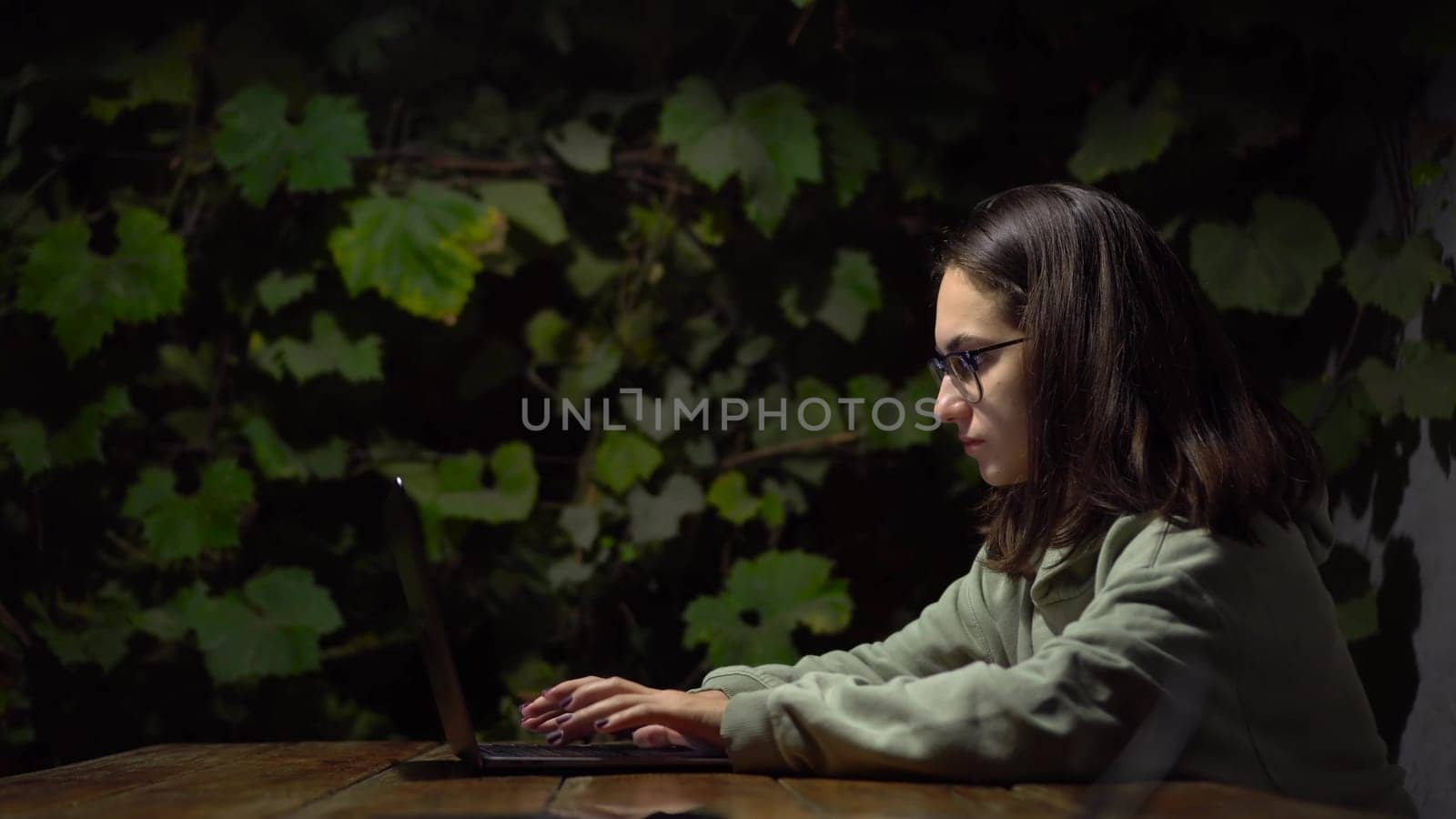 Young woman at a table in a gazebo typing on a laptop late at night. A girl in a hoodie and glasses with a laptop on a background of green grapes. 4k