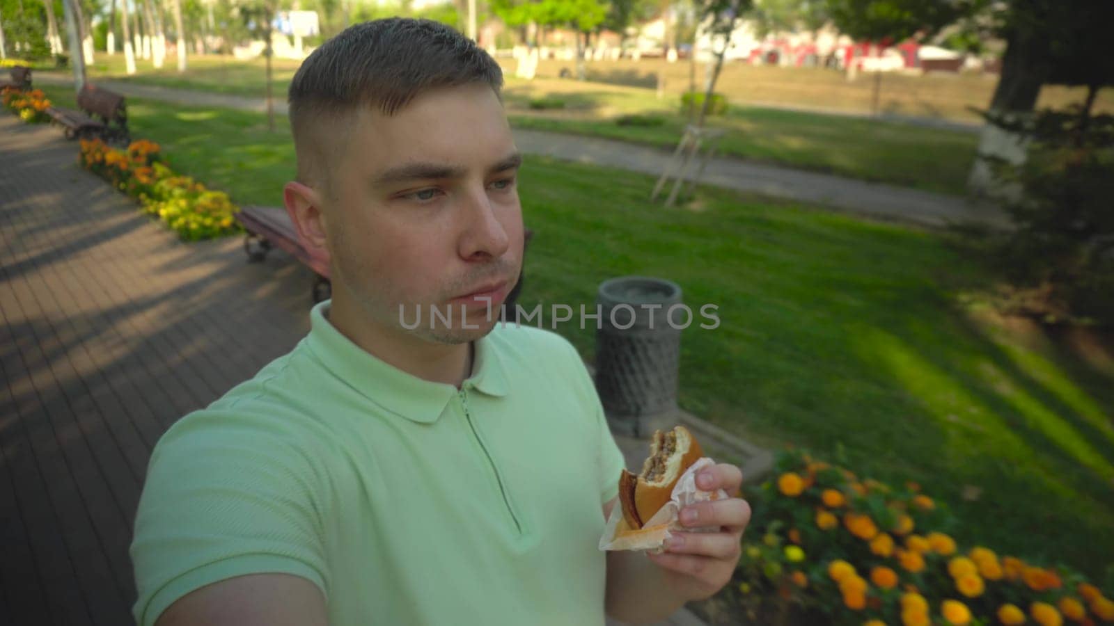 A young man walks through the park and eats a burger. A man films himself while walking along a green alley. 4k