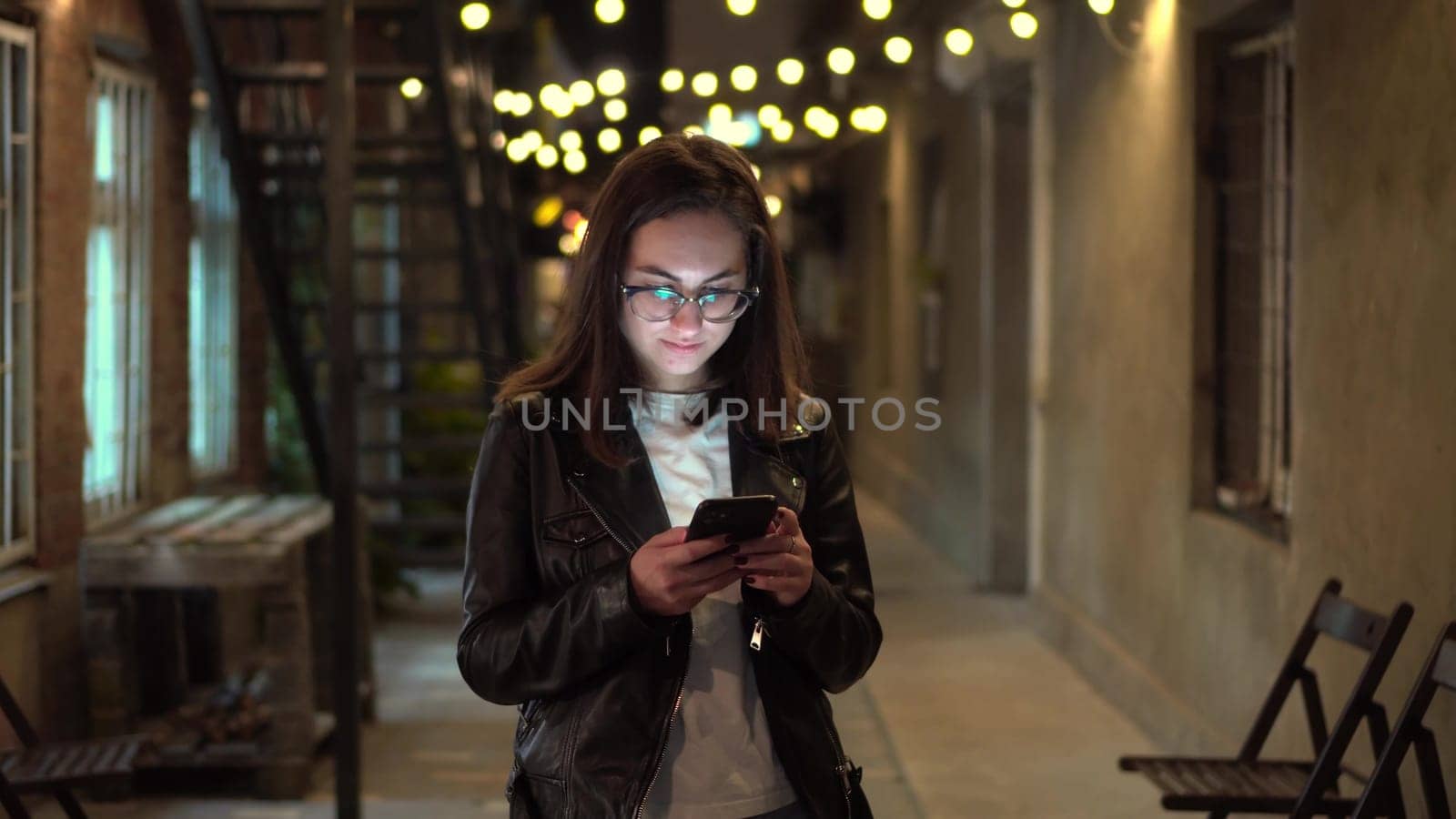 A young woman texts on the phone on a narrow street in the evening. A girl in glasses and a leather jacket on a cozy street with a phone in her hands. 4k