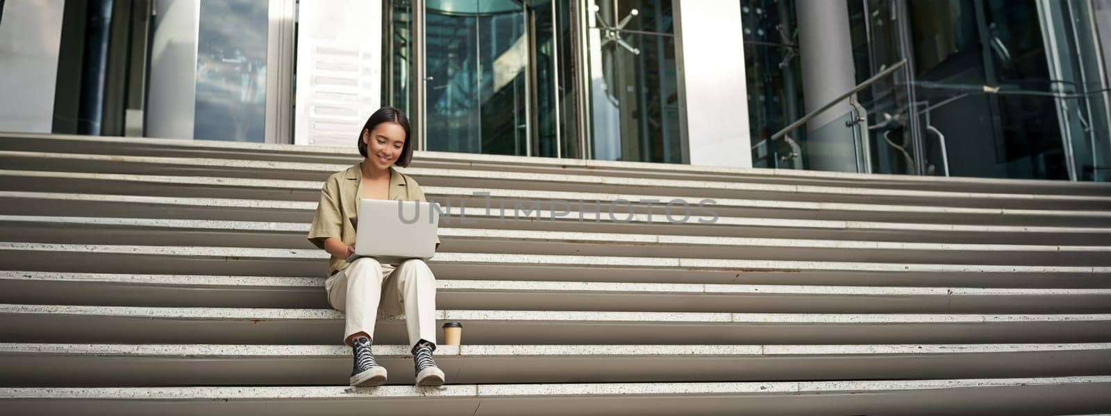 Asian girl student sits on stairs near campus, types on laptop, does her homework outdoors by Benzoix