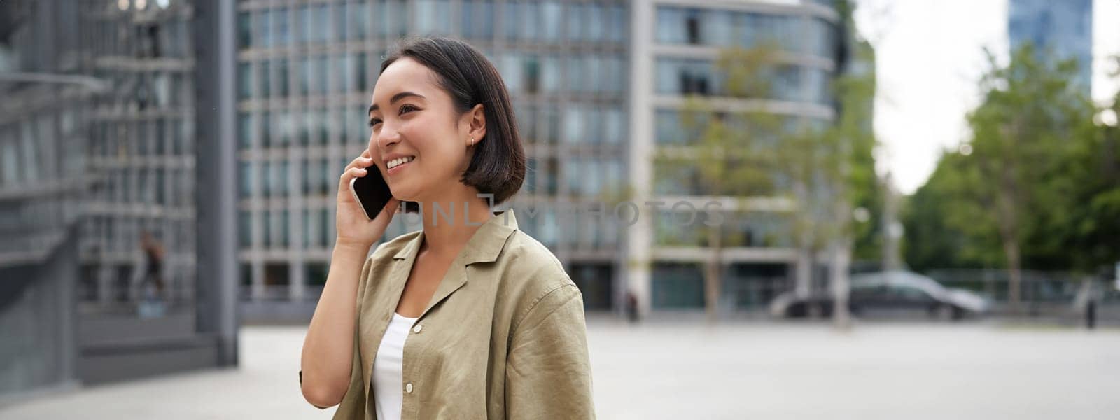 Vertical shot of young asian digital nomad, girl talks on mobile phone and walks on street with laptop. Young woman remote worker going to coworking space by Benzoix