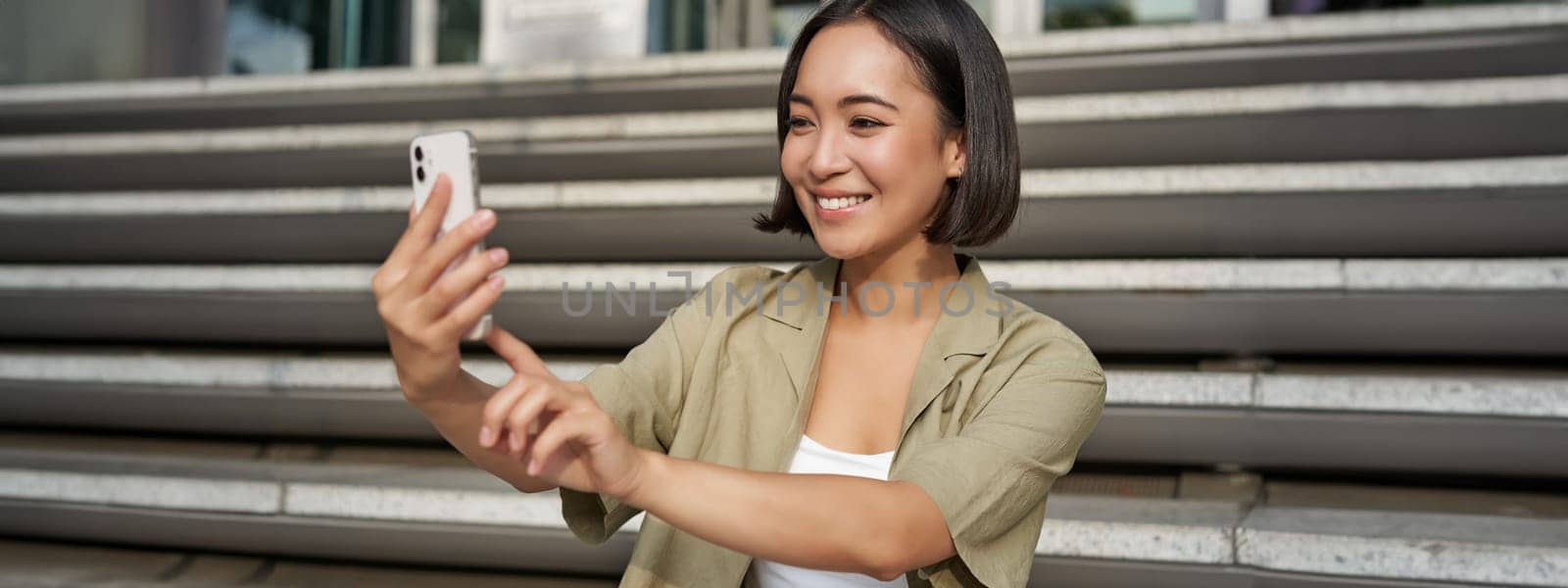 Portrait of asian beautiful girl takes selfie on smartphone. Young korean woman sits on stairs outdoors and makes photos on mobile phone by Benzoix