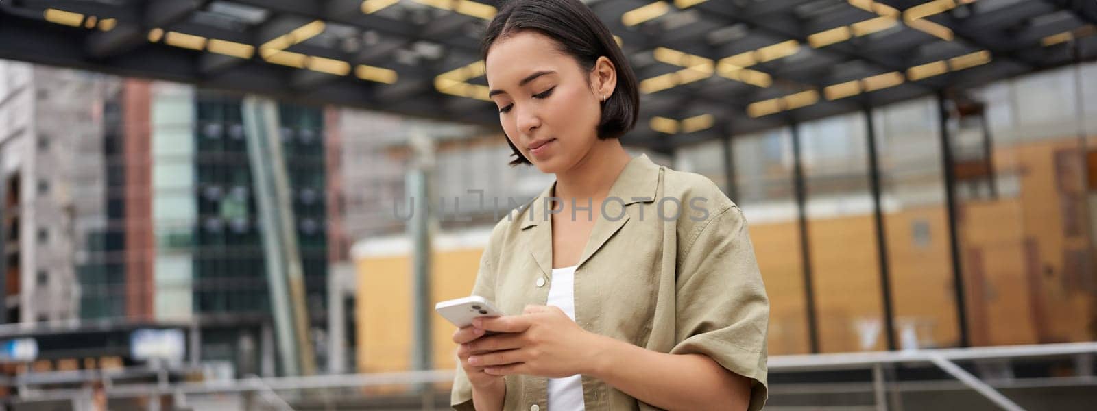 Technology people. Young smiling asian girl holding smartphone, using mobile app while standing on street, navigating around city.