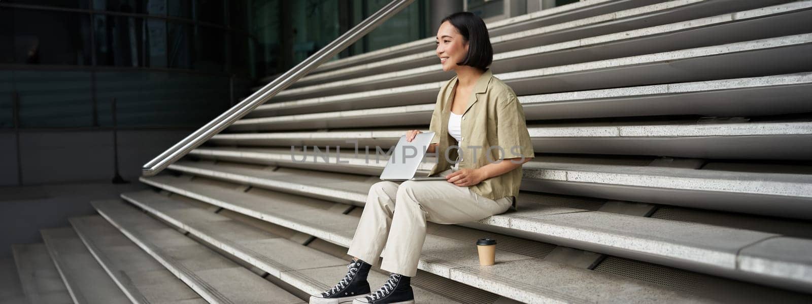 Young smiling asian girl with laptop, sits on street of city with coffee. Young woman does homework on computer while sitting on street stairs, digital nomad works remote by Benzoix