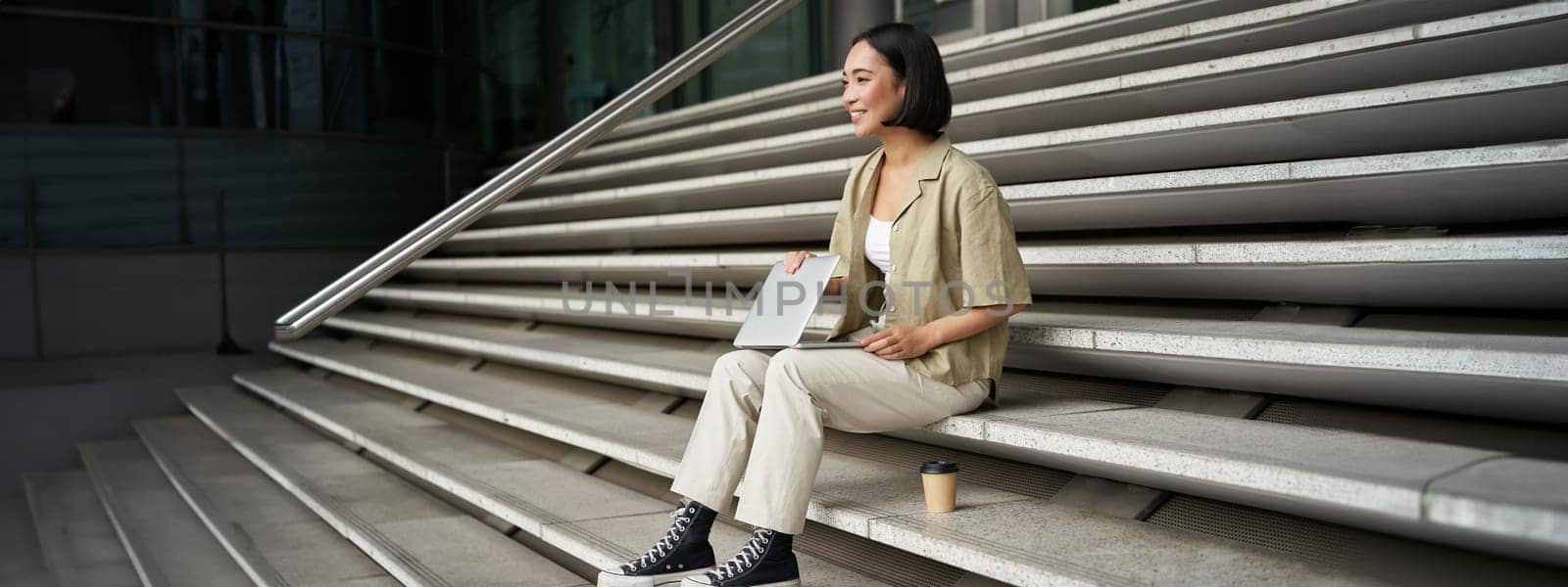 Young smiling asian girl with laptop, sits on street of city with coffee. Young woman does homework on computer while sitting on street stairs, digital nomad works remote.