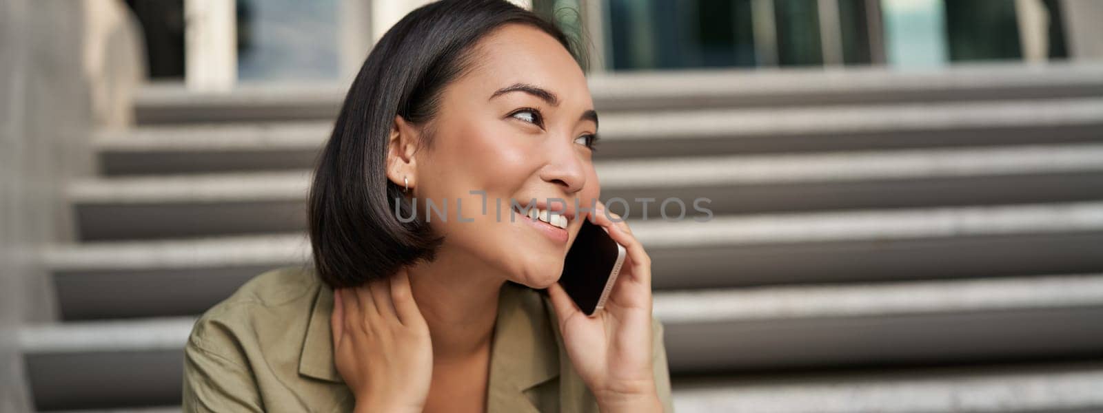 Cellular technology. Smiling asian girl talks on mobile phone, makes a call. Young woman with telephone sits on stairs by Benzoix