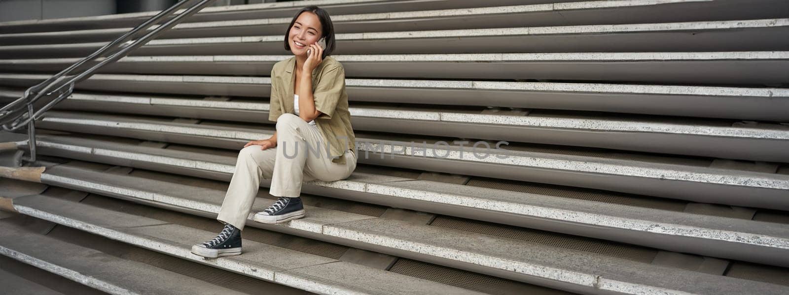 Portrait of asian student girl on stairs talks on mobile phone, smiles at camera, sits outside building by Benzoix