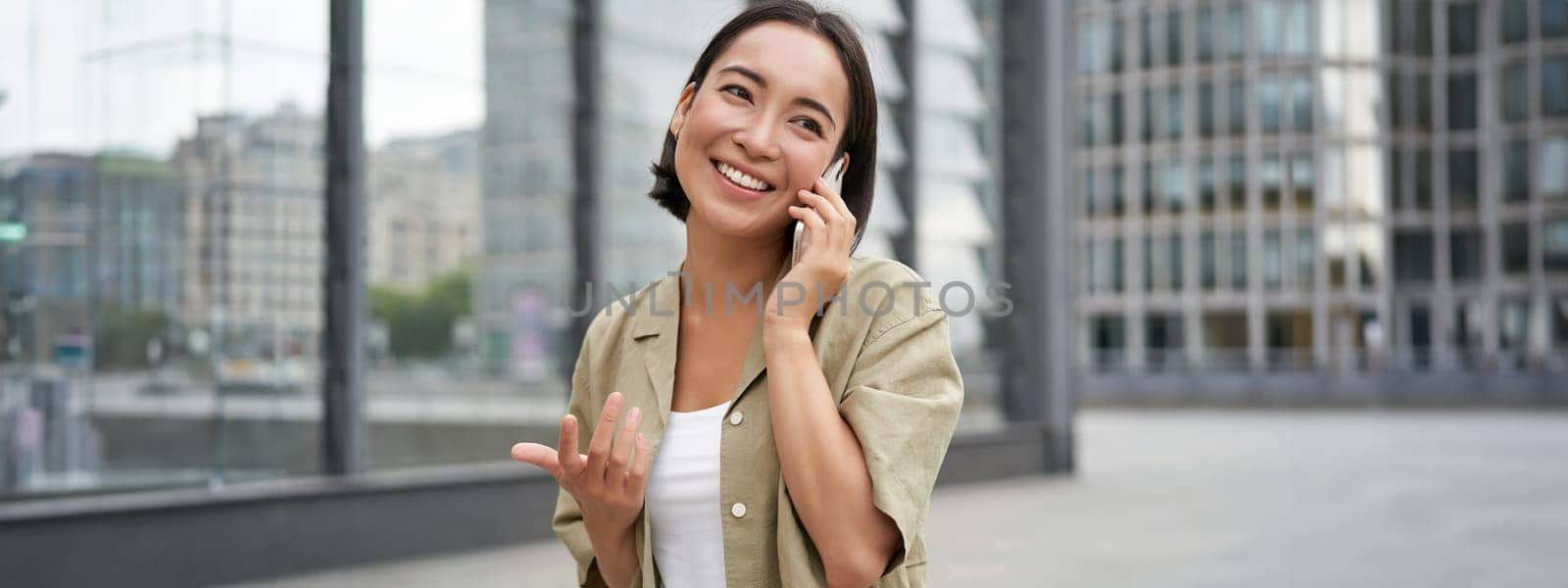 Cellular connection. Young asian woman makes a telephone call, talking on mobile smartphone and walking on street.