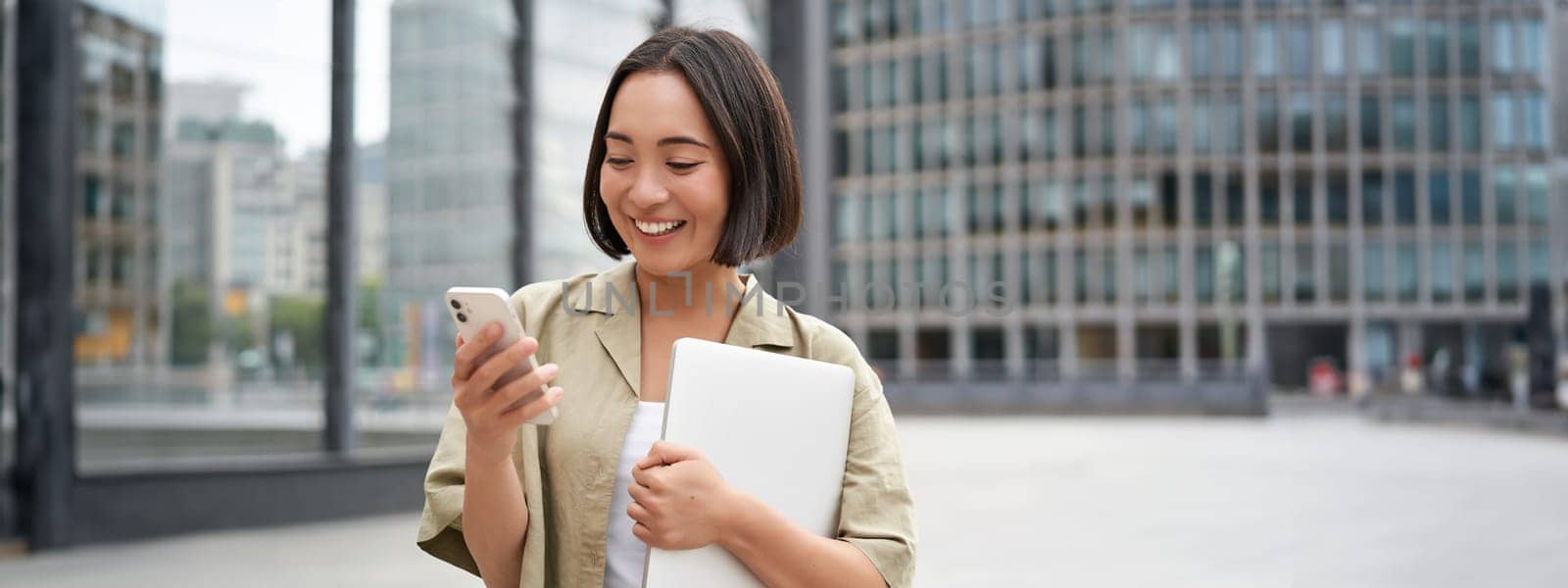 Portrait of young smiling asian woman walking on street, going to work with laptop and smartphone by Benzoix