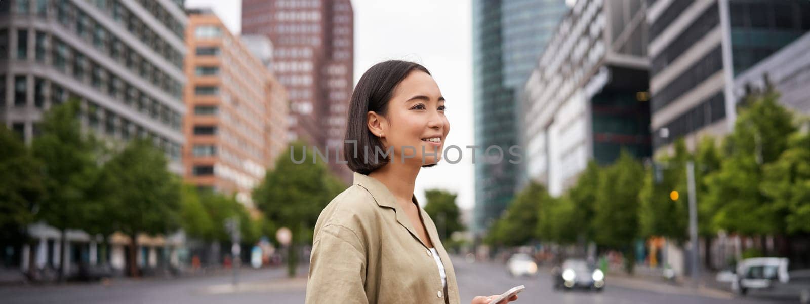 Young asian woman exploring city with smartphone app, holding mobile phone and walking on screet.