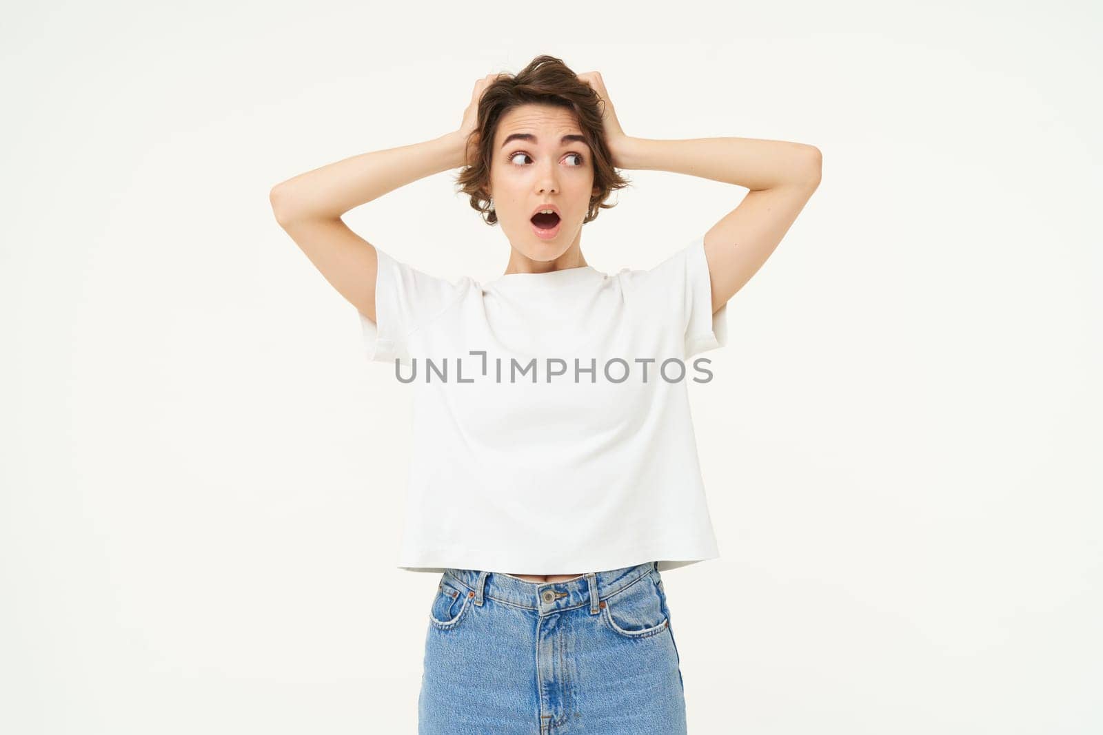Portrait of girl in panic, grabs her head with both hands, looking shocked, worried about something, standing over white background.