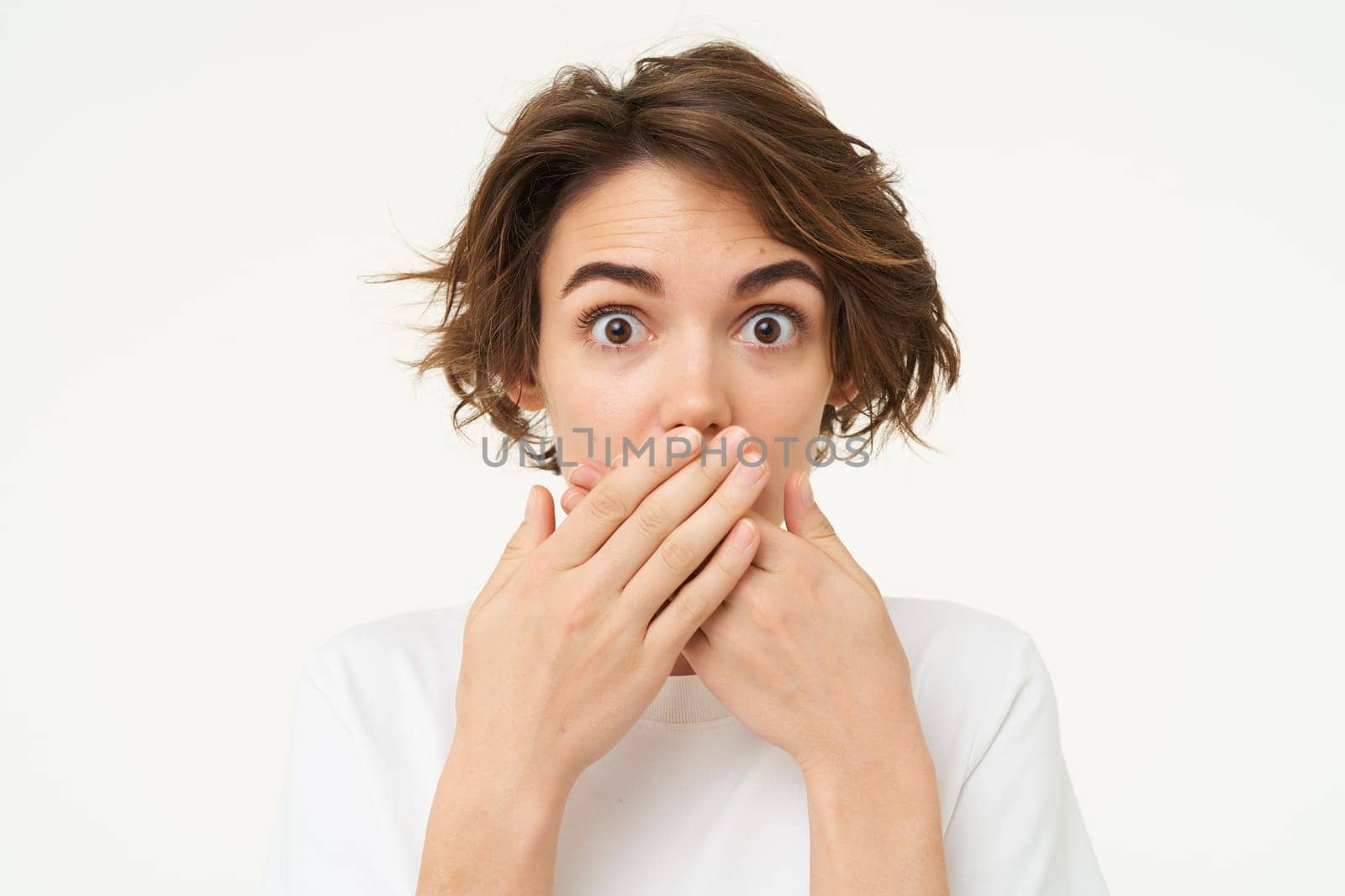 Image of brunette woman shuts her mouth, looks surprised, puts hands over her lips, stands over white background.