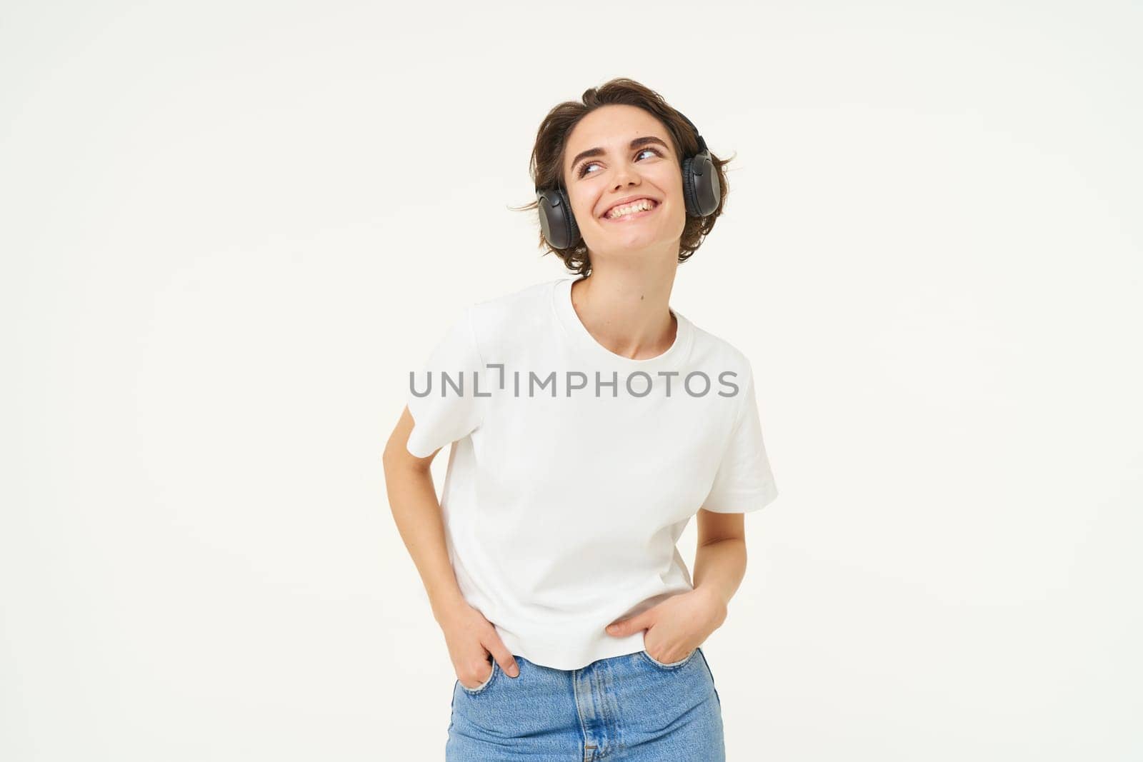 Image of modern girl in wireless headphones, listens to music in earphones, smiles and looks happy, poses in casual clothes over white background.