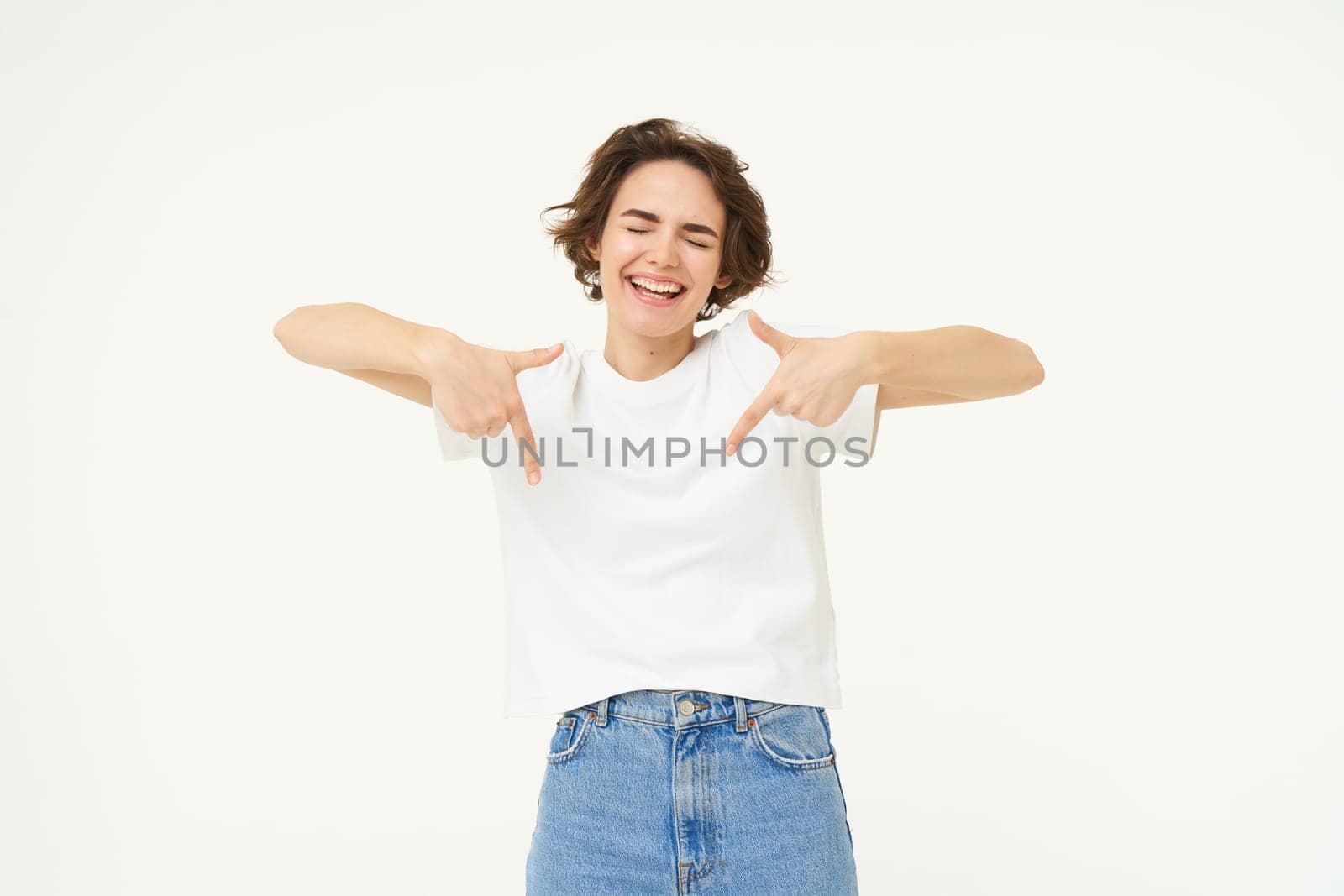 Image of beautiful, happy brunette woman, pointing fingers down, showing advertisement, standing over white studio background by Benzoix