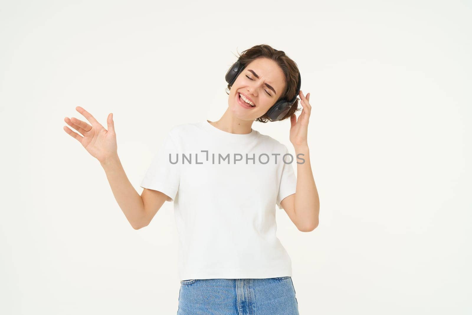 Happy girl dancing and having fun, listens to music in wireless headphones, stands over white background.