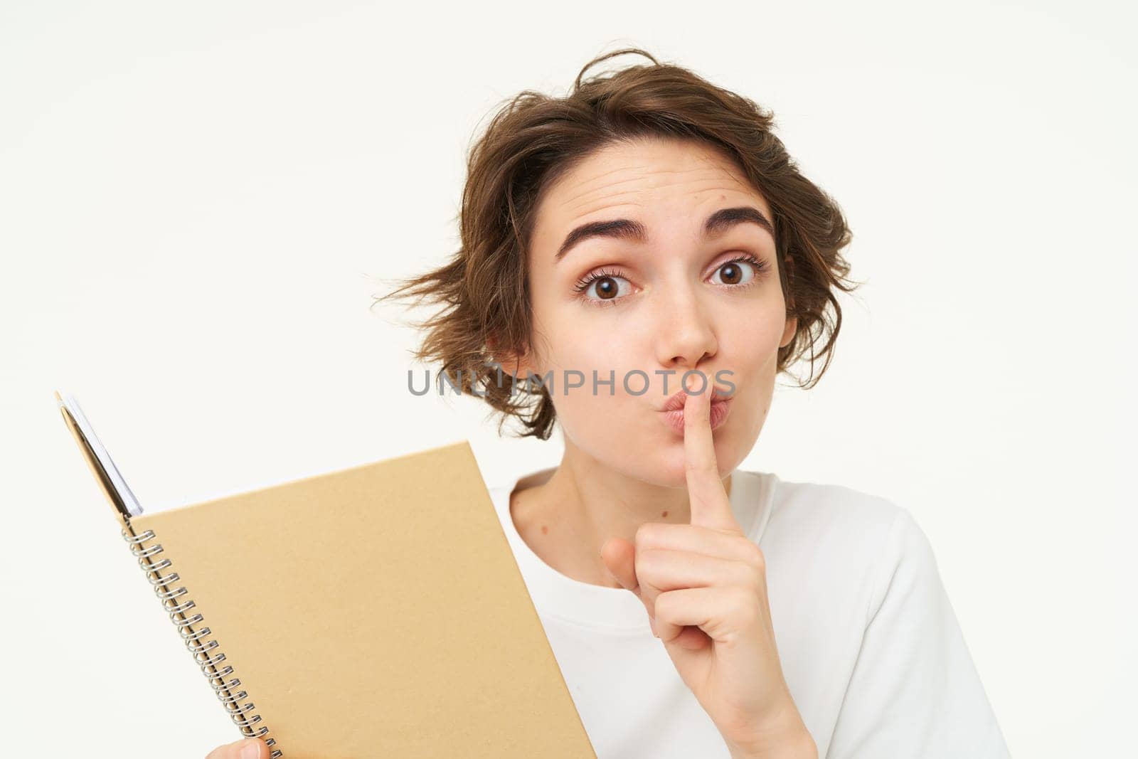 Young woman reads her diary, makes shhh shush face, press finger to lips, hiding something, asking to keep quiet, save secret, standing over white background by Benzoix