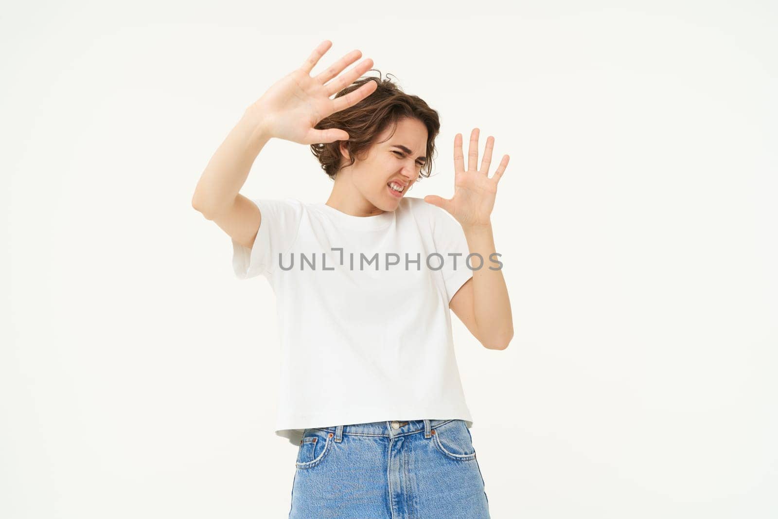 Portrait of woman raising hands in defense and blocking face from something, standing over white background.
