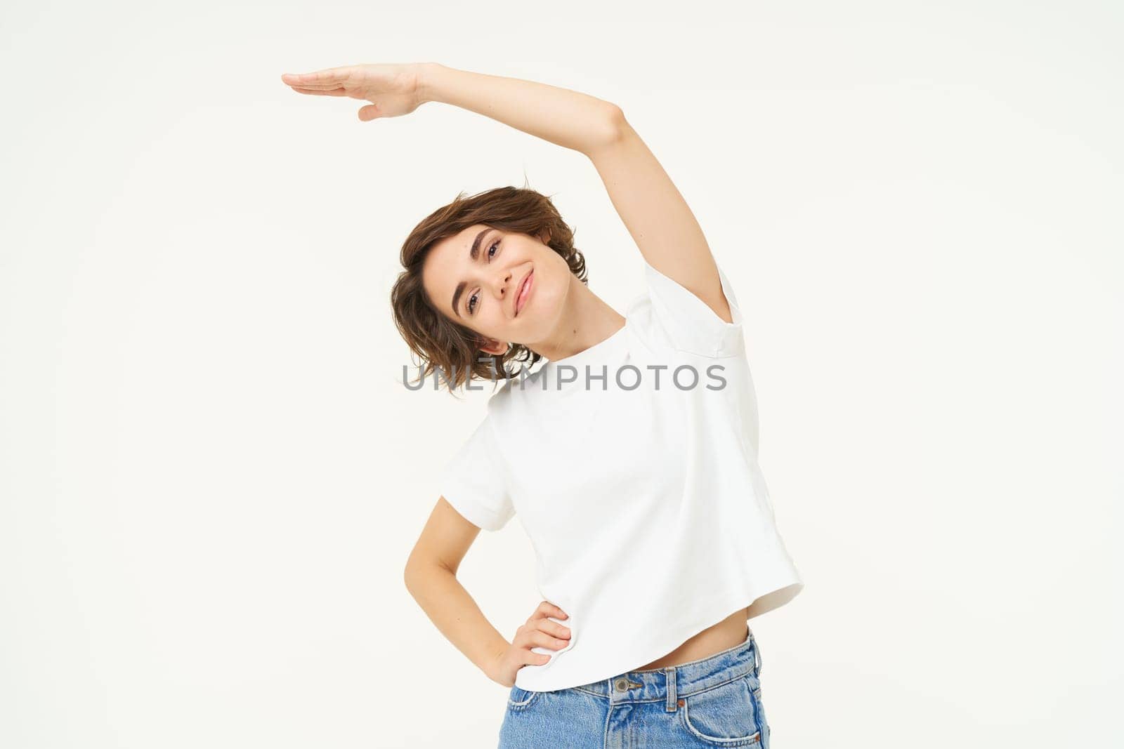 Portrait of woman doing exercise, fitness movements, bending arm sideways and stretching body, standing over white background. Copy space