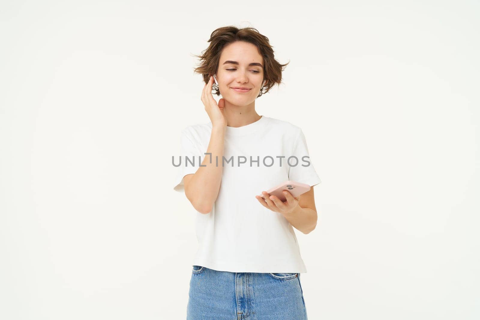Portrait of woman puts on wireless headphones and holds smartphone, listens music, stands over white background.