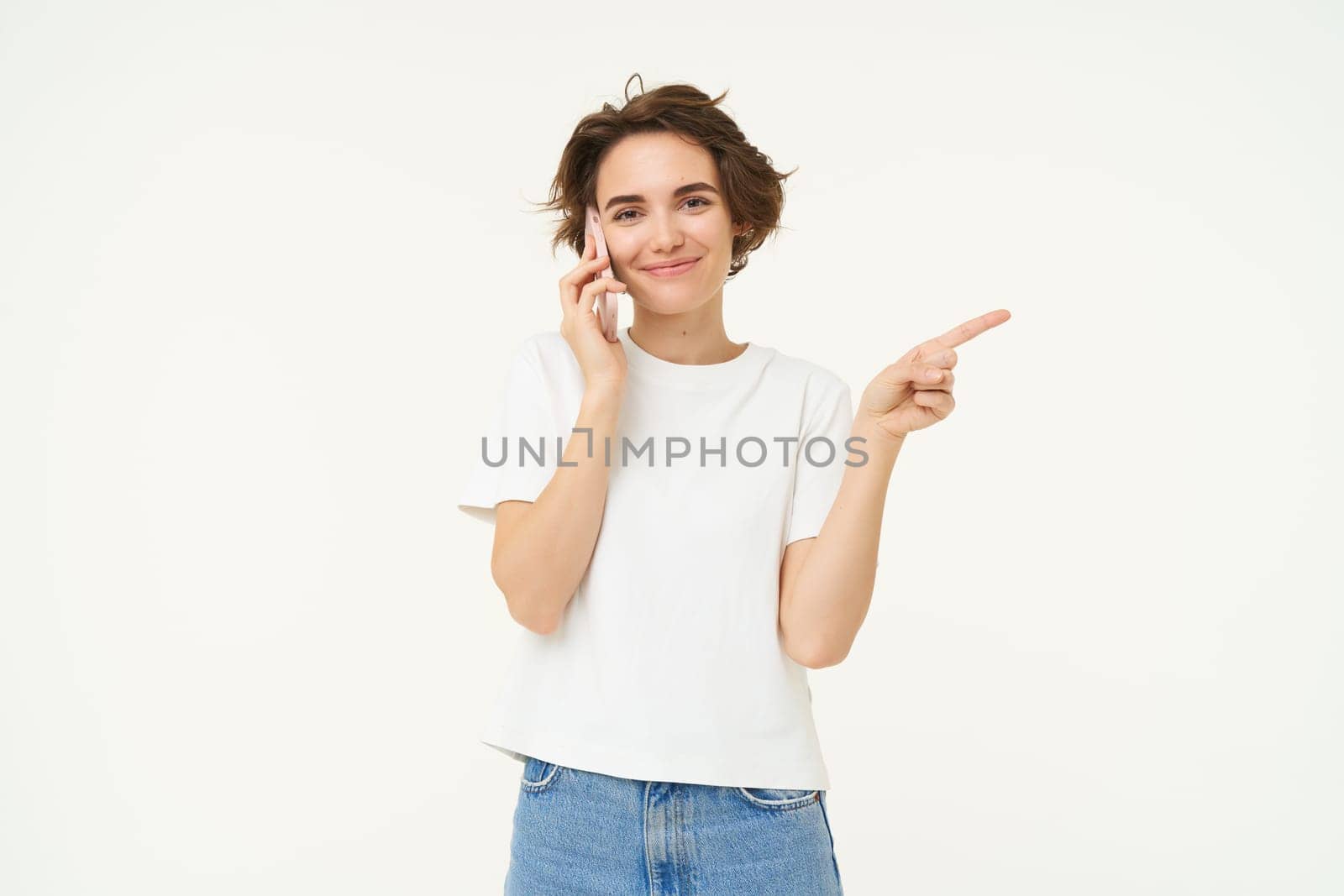 Portrait of young woman talking on mobile phone, using smartphone and pointing finger right, showing direction, banner advertisement, studio background.