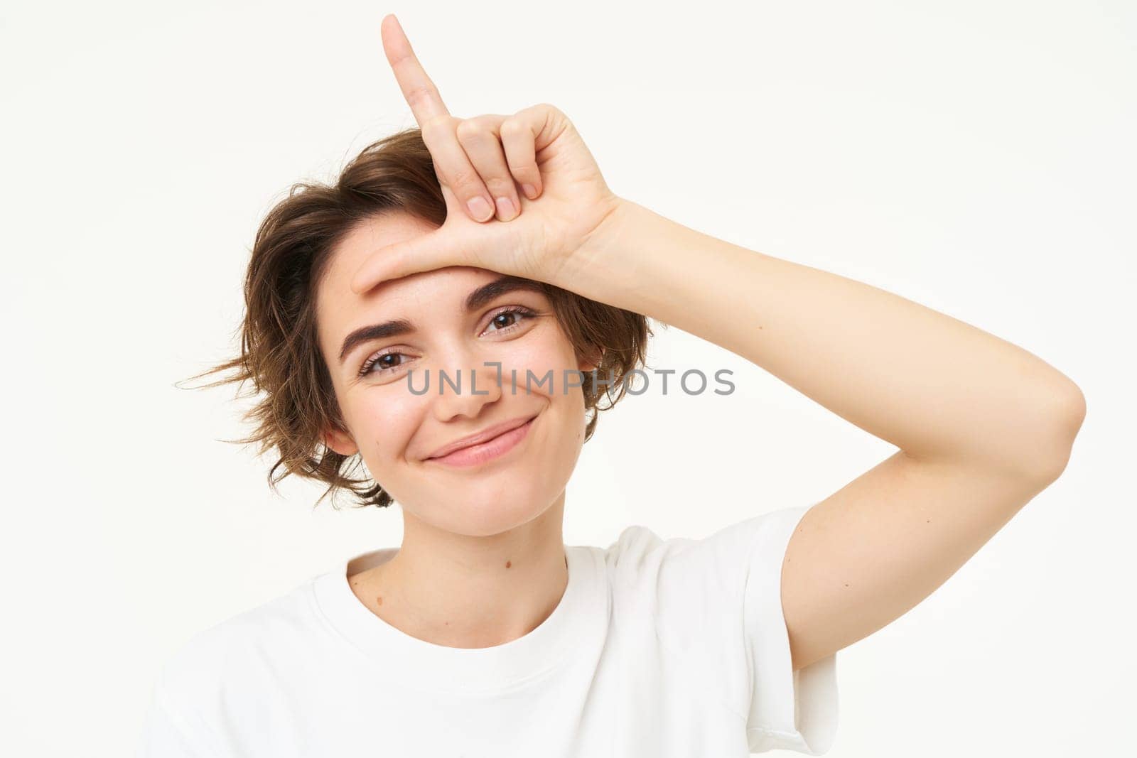 Close up of young woman making fun of friend, smiling and showing loser gesture, l letter on forehead, standing over white background.