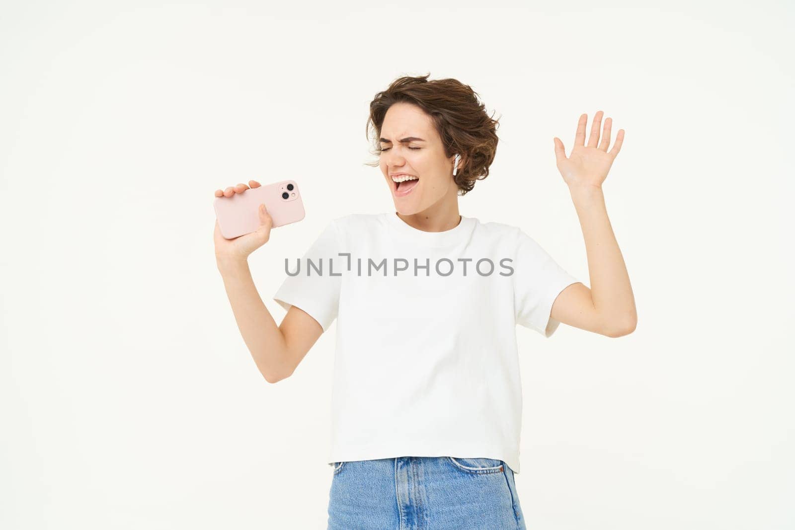 Portrait of carefree girl singing in wireless headphones into smartphone microphone, playing music game on mobile app, standing over white background by Benzoix