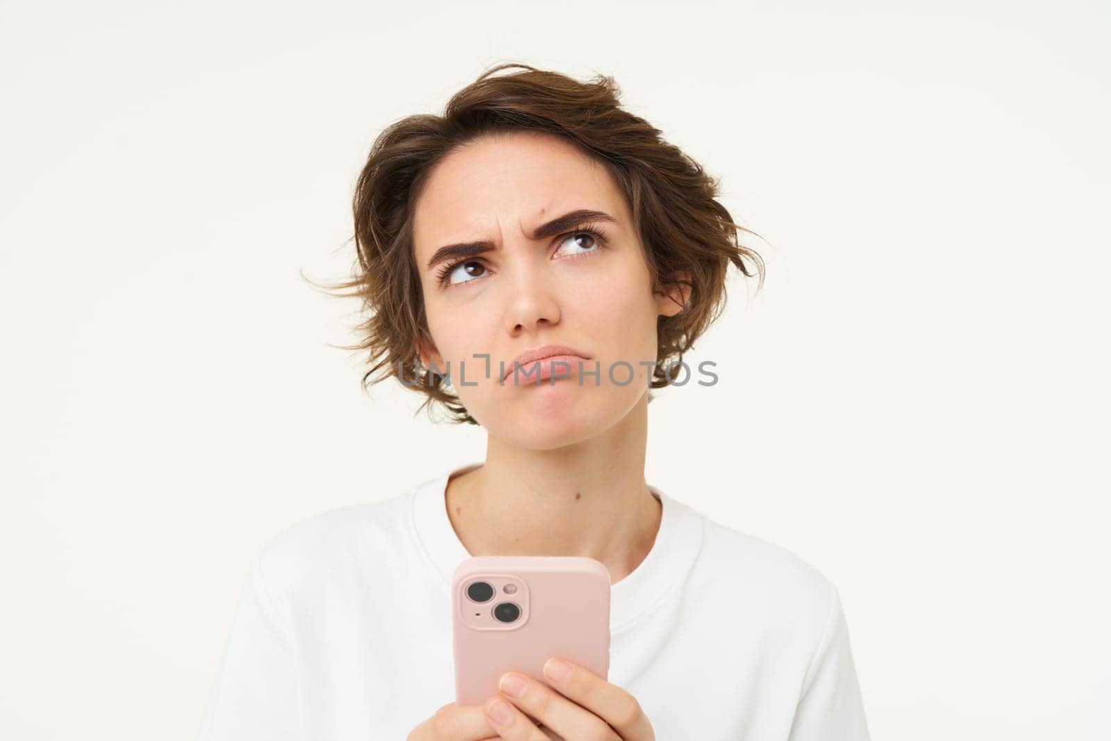 Portrait of puzzled brunette woman, standing with smartphone, thinking and frowning, looking reluctant, standing over white background.